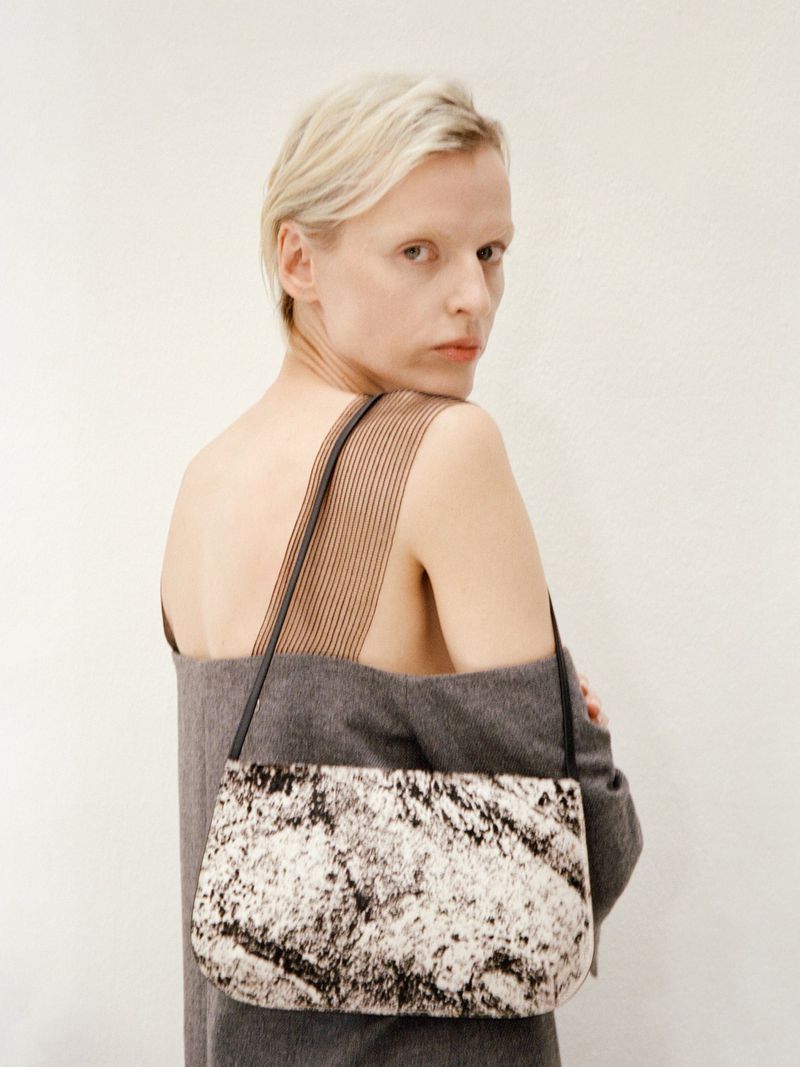 Blonde woman wearing a grey off-the-shoulder top carrying a patterned shoulder bag against a plain white studio background.