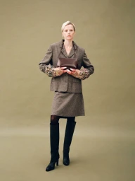 Model in a brown tweed suit holding a dark leather handbag against a solid olive green studio backdrop.