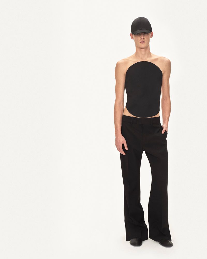 Studio shot of an adult male in a black round top and flared black pants against a white backdrop, striking a fashion pose.
