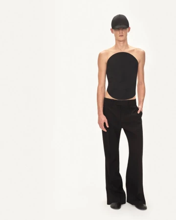 Studio shot of an adult male in a black round top and flared black pants against a white backdrop, striking a fashion pose.