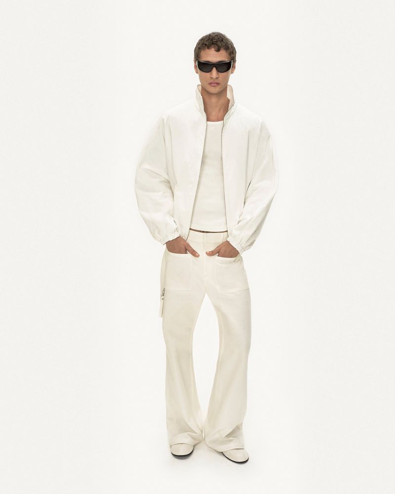 Full-length studio shot of a slender adult male wearing a white jacket, white tank top, wide-leg pants, sunglasses, and matching shoes.