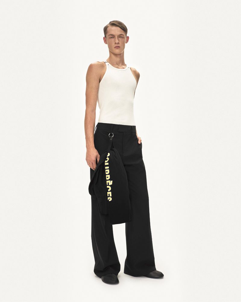 Full-length studio shot of a young adult male fashion model wearing a white tank top and black wide-leg pants, holding a black bag.