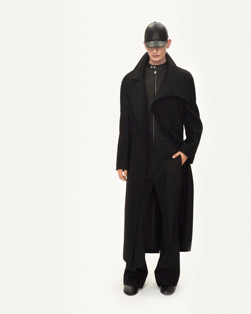 Studio shot of adult male model in long black coat, black trousers, and leather baseball cap against a plain white backdrop. Fashion.