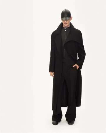 Studio shot of adult male model in long black coat, black trousers, and leather baseball cap against a plain white backdrop. Fashion.