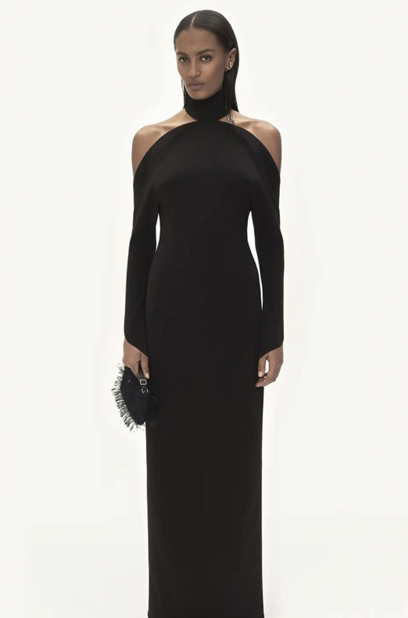 Elegant woman in a long black gown with open shoulders, posing against a clean white backdrop, holding a small round handbag.