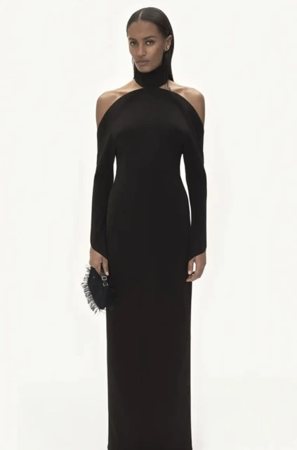 Elegant woman in a long black gown with open shoulders, posing against a clean white backdrop, holding a small round handbag.