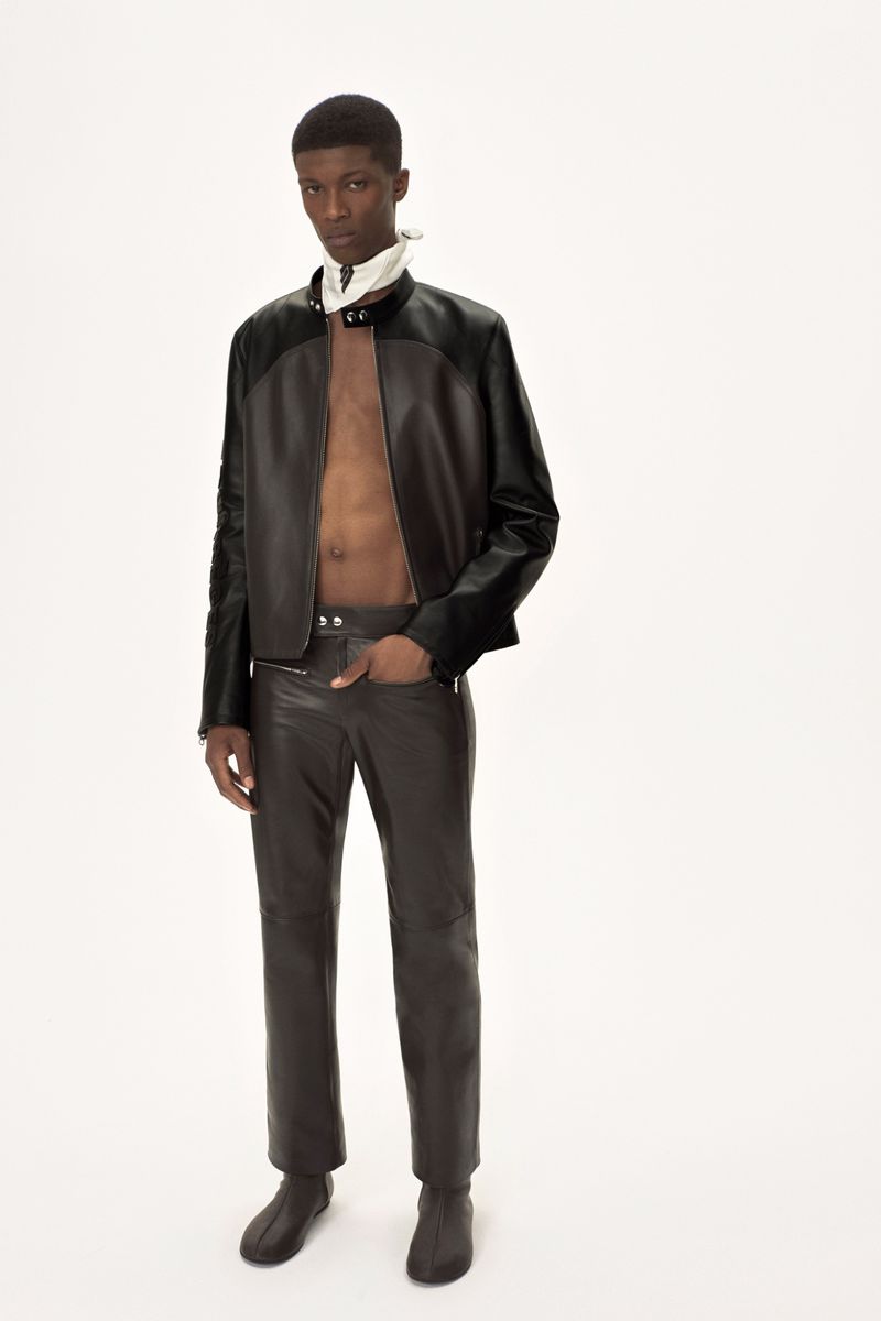 Fashion shot of African American male model in brown leather jacket and pants posing against a white backdrop in a photo studio.