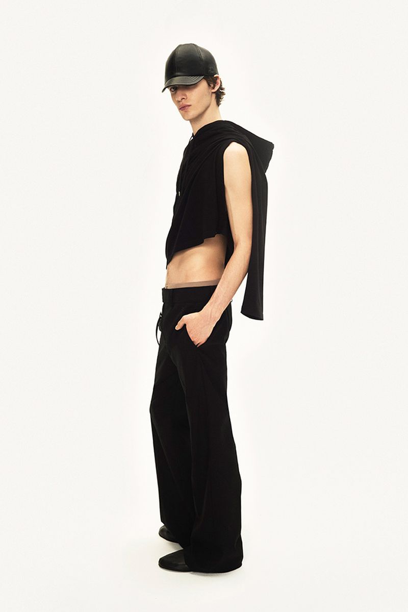 Studio shot of a slim male model in black cap, sleeveless hoodie, and wide-legged pants striking a fashion pose against white backdrop.