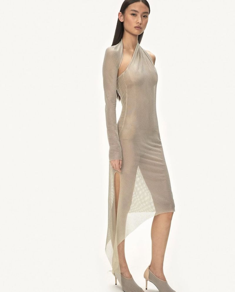 Elegant woman in a tan, sheer, halter-style dress with a thigh-high slit, accessorized with neutral heels against a bright white background.