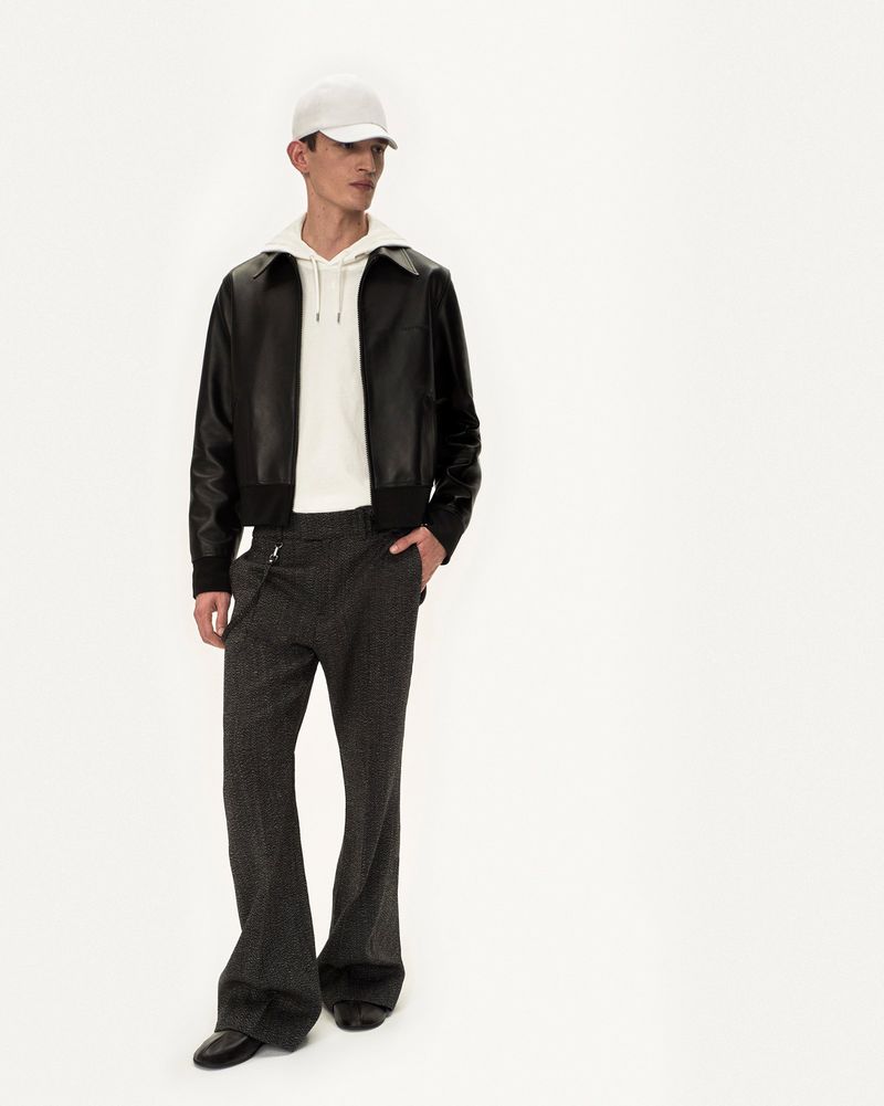Male fashion model in leather jacket, hoodie, trousers, cap and dress shoes posing in bright photo studio against white backdrop.