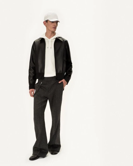 Male fashion model in leather jacket, hoodie, trousers, cap and dress shoes posing in bright photo studio against white backdrop.