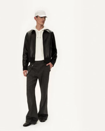 Male fashion model in leather jacket, hoodie, trousers, cap and dress shoes posing in bright photo studio against white backdrop.