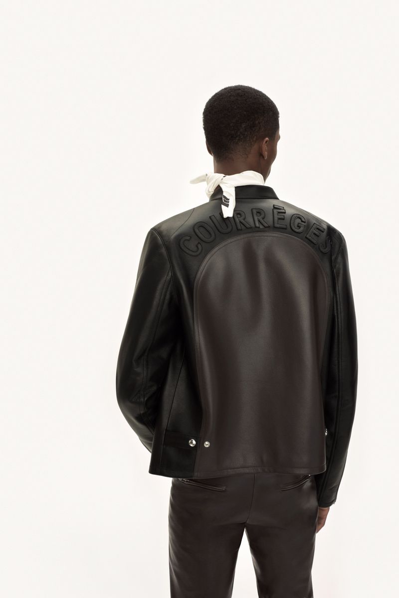 Model seen from behind wearing a dark leather jacket with embossed lettering against a seamless white background.
