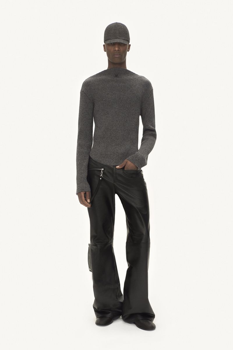 A person in a gray cap and sweater, accessorized with wide-leg leather pants and dress shoes, all set against a stark white backdrop.