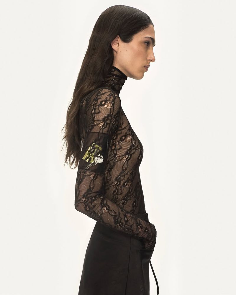 Fashionable adult female in a side-view studio shot, wearing a black lace turtleneck top, with black bottoms and long dark hair.