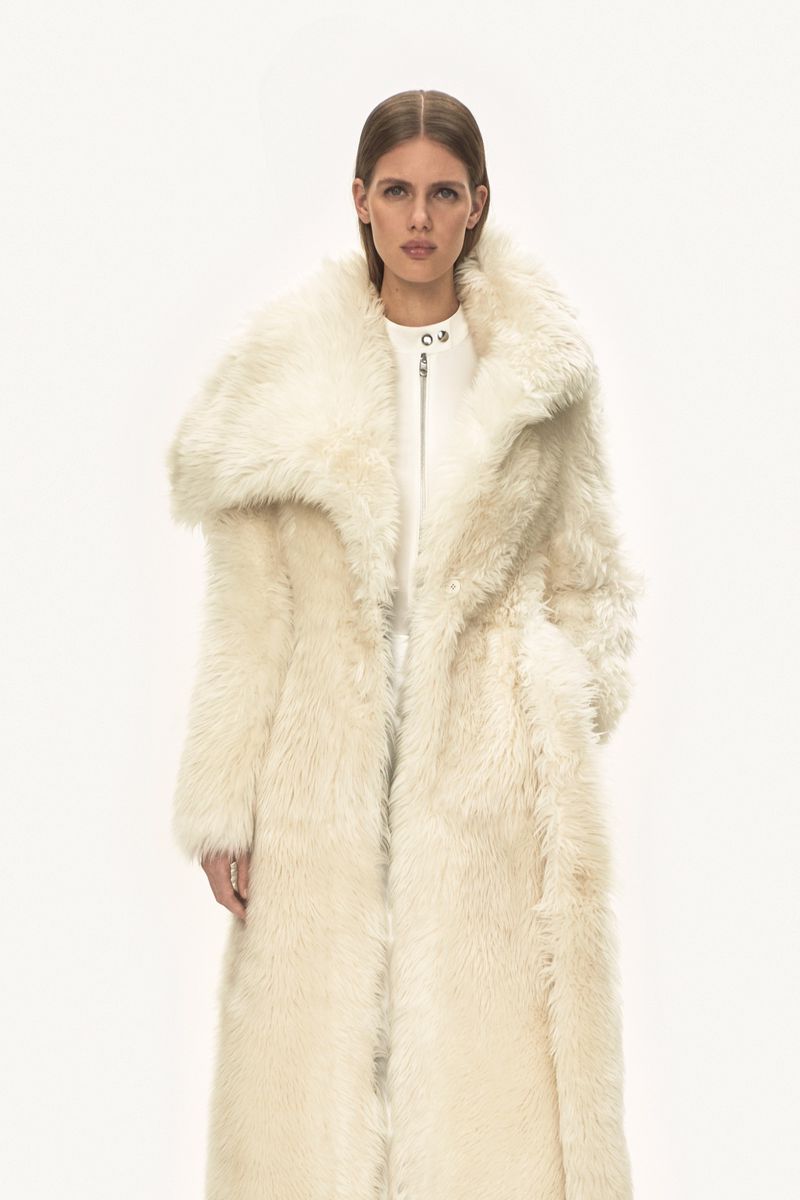 A woman poses in a long, oversized, cream-colored faux fur coat against a white background in a studio shot for fashion editorial.