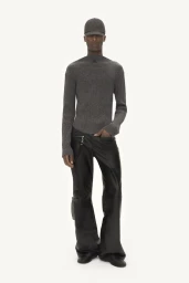 Model wearing a grey knit sweater, black leather trousers, and a grey cap against a white studio background.