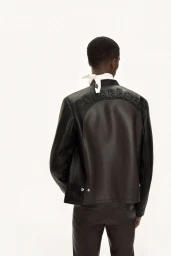 Model seen from behind wearing a dark leather jacket with embossed lettering against a seamless white background.