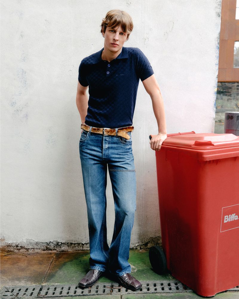 Full-length portrait of a male model in a navy polo shirt and blue jeans, leaning against a red recycling bin on an urban street.