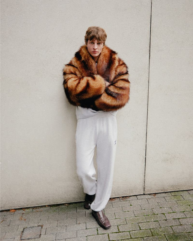 Adult male model in a fur coat and sweatpants poses against a wall in a medium shot with natural lighting outdoors.