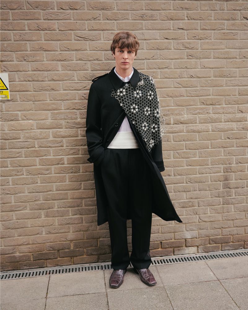 A fashionable adult male with brown hair wearing a long black trench coat and dress shoes poses against a brick wall in an outdoor urban setting.