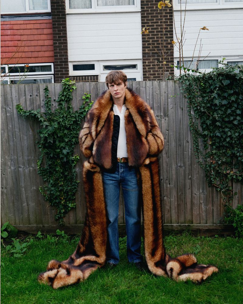Man poses in a brown fur coat with a long train in a backyard with grass, a wooden fence, a brick building, and green foliage.