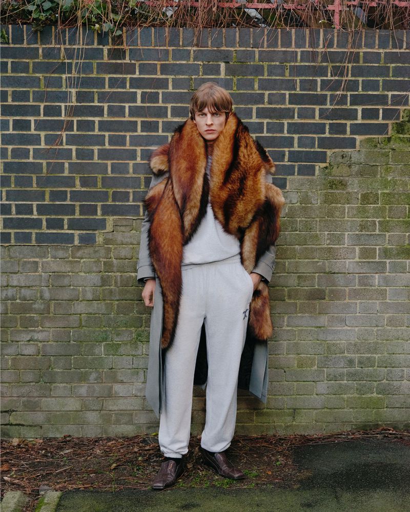 Man in a light gray tracksuit and brown shoes stands against a brick wall, wearing a large brown fur stole and a long gray coat.