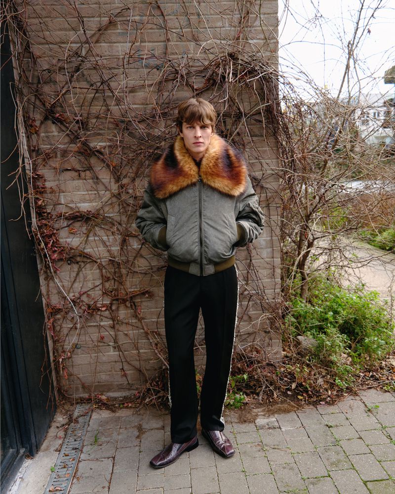 Full-length portrait of a young man in an olive bomber jacket with fur collar and black pants with white stripe standing outdoors.
