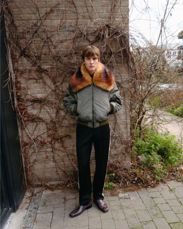 Full-length portrait of a young man in an olive bomber jacket with fur collar and black pants with white stripe standing outdoors.