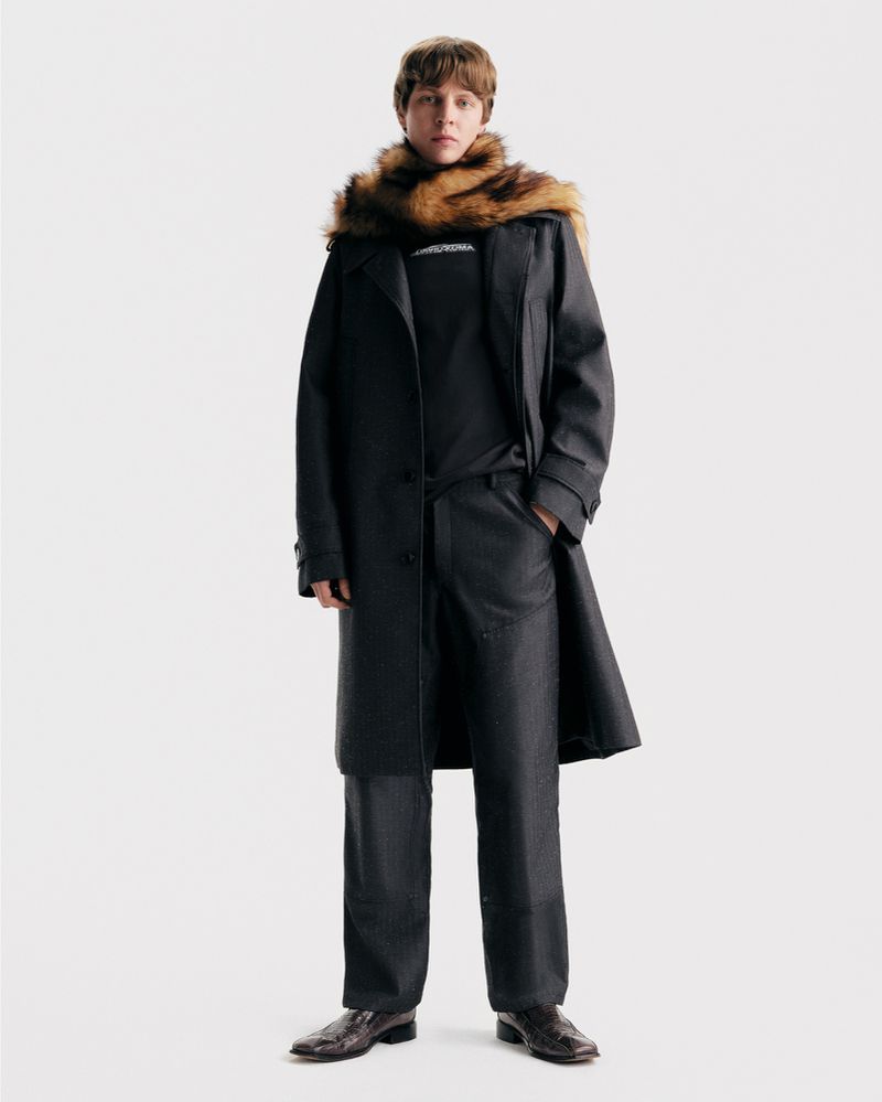 Full-length studio shot of a person in a dark coat, trousers, and fur stole, standing against a white backdrop, showcasing fashion and style.