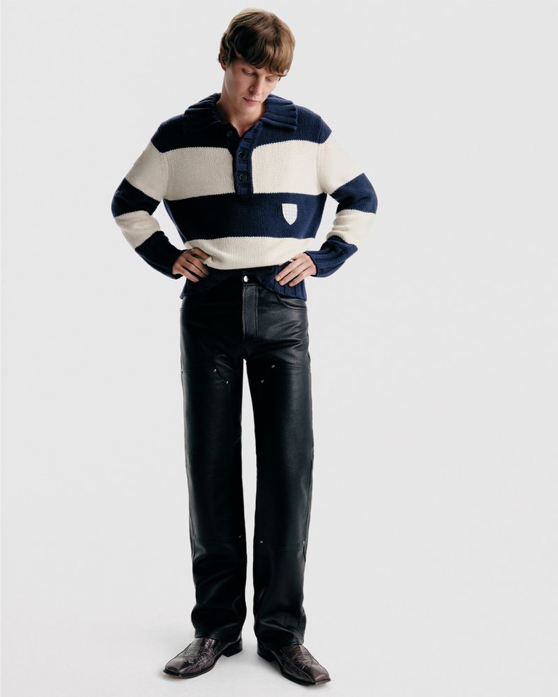 Full-length shot of an adult man wearing a striped sweater and leather pants, standing against a neutral backdrop in a studio setting.