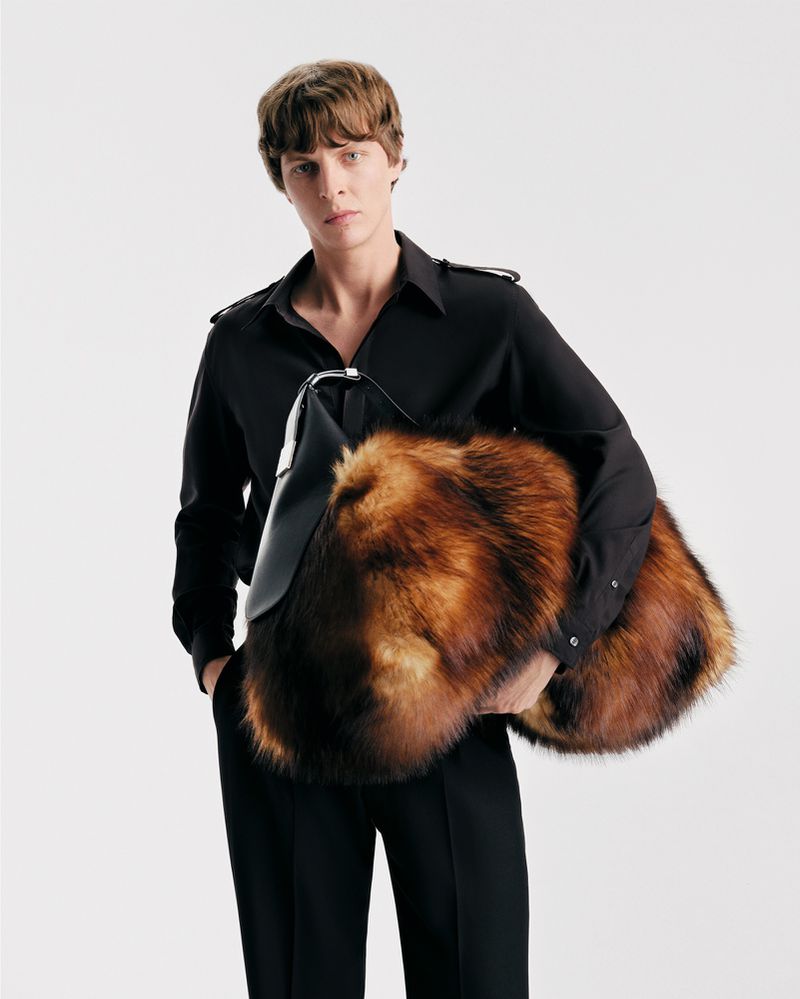 Studio shot of a male fashion model in a black outfit holding a fur-covered bag, set against a clean white backdrop. Fashion and style.