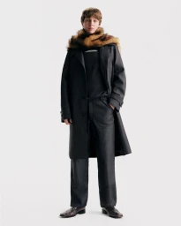 Model wearing a dark long coat, black trousers, and a furry scarf against a white background
