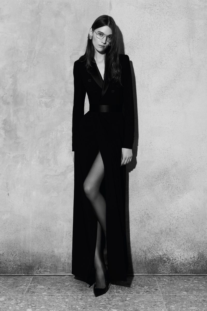 Elegant woman posing in a floor-length black dress with a slit and heels against a textured gray background in a fashion portrait.