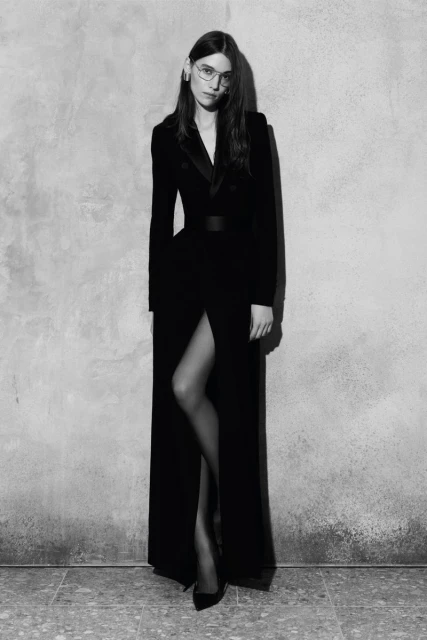 Elegant woman posing in a floor-length black dress with a slit and heels against a textured gray background in a fashion portrait.