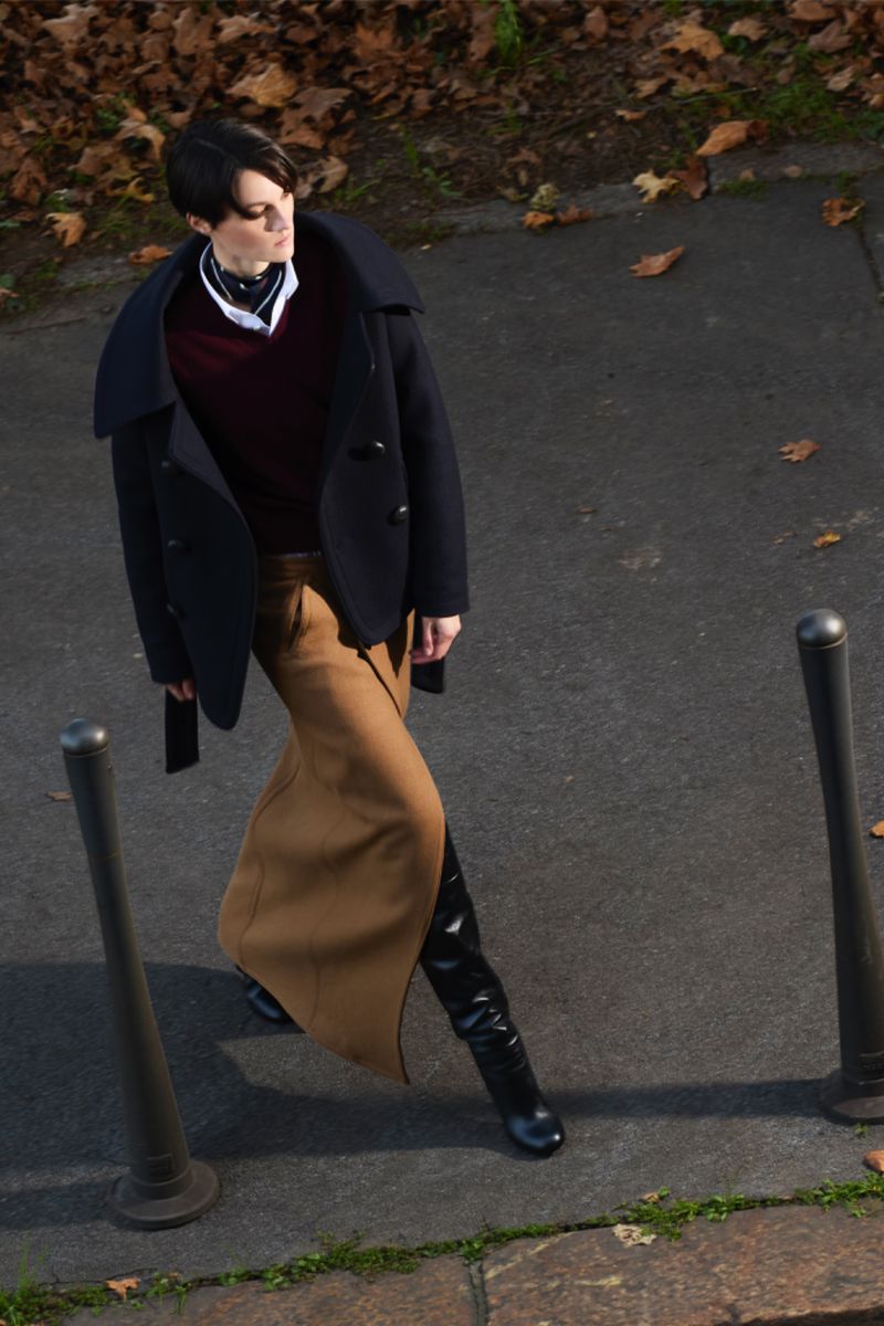 High-angle view of a stylish woman in a coat and skirt walking on a paved surface with autumn leaves, captured in natural light.