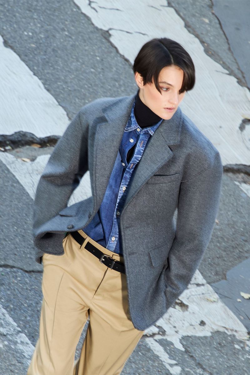 Adult female model wearing a gray blazer, blue collared shirt, black turtleneck, tan pants, and black belt on a city crosswalk.