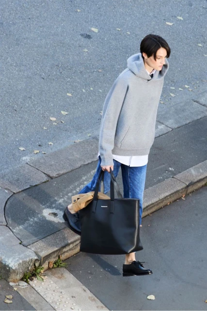 Woman in gray hoodie and jeans walks on the street, holding a black tote bag in daylight, captured from a high angle view.