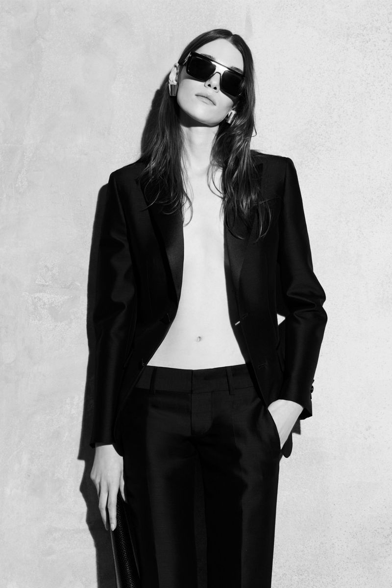 Model wearing a black blazer and trousers with dark sunglasses against a textured wall in a black and white portrait.