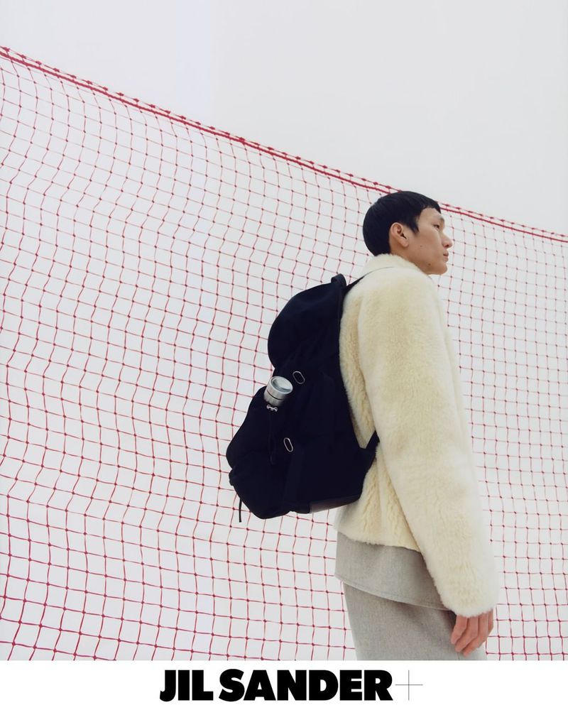 Asian male model wears fur jacket and backpack against red net background for JIL SANDER+ fashion editorial, outdoors.