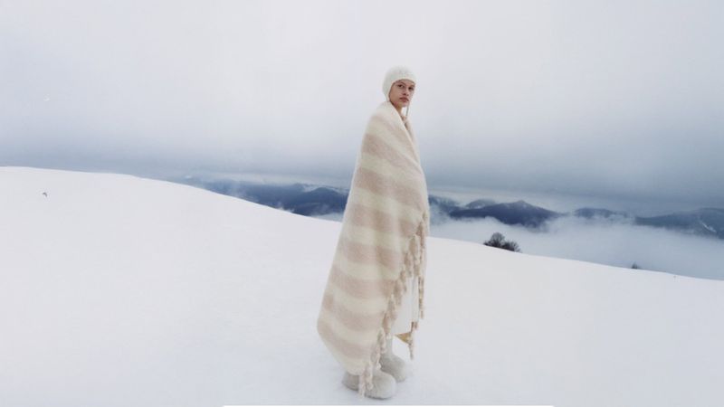 Model wrapped in a striped wool blanket and wearing a matching beanie standing on a snowy mountain ridge.