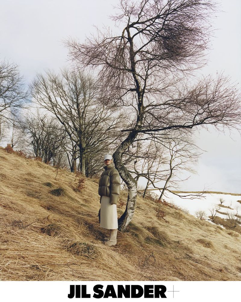 A woman in an olive puffer jacket, cream skirt, and hat poses on a hillside with bare trees, in a fashion editorial style image.