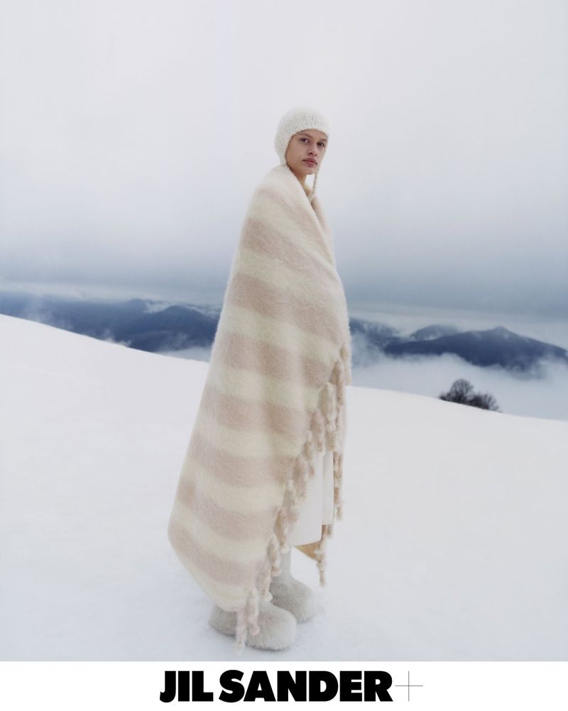 Fashion shot of an adult female wearing a white knit hat and draped in a cream striped blanket in a snowy landscape; Jil Sander logo.