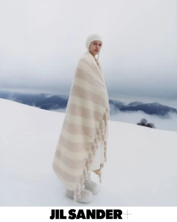 Fashion shot of an adult female wearing a white knit hat and draped in a cream striped blanket in a snowy landscape; Jil Sander logo.