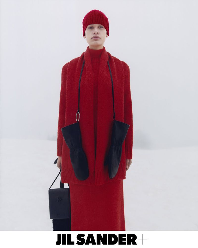 A person in a red knit outfit holds a black bag in a studio setting, standing against a bright background wearing mittens.