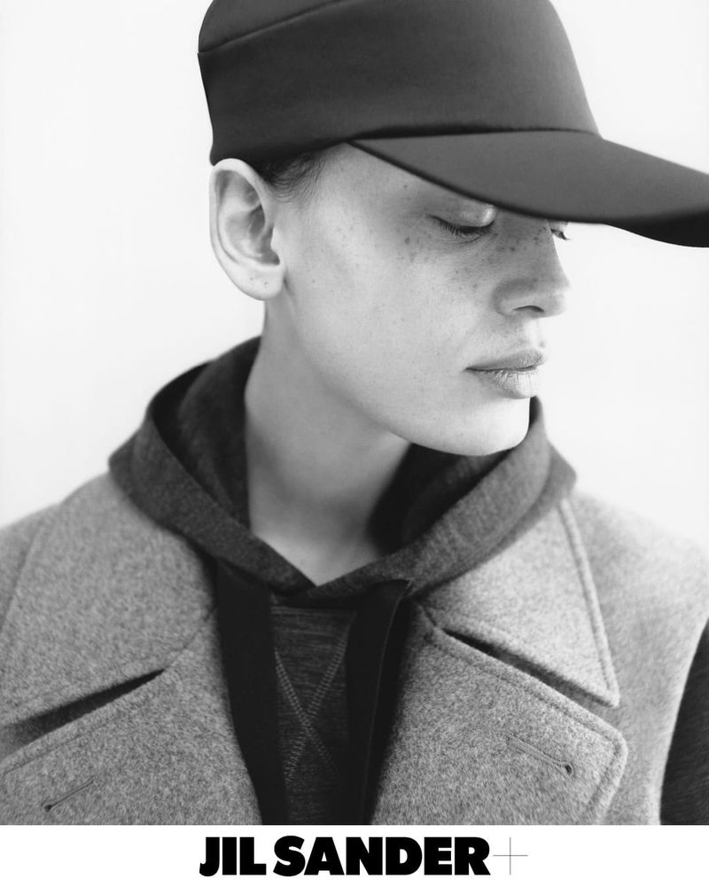 Close-up black and white portrait of a model wearing a dark cap and wool coat for Jil Sander+ Fall 2024 campaign.