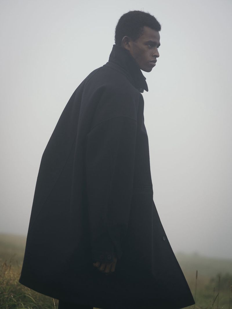 Dark-skinned male model in a black overcoat walking through grassy fog-covered terrain, fashion editorial style, medium shot, natural light.