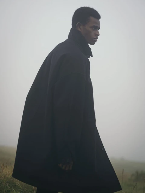 Dark-skinned male model in a black overcoat walking through grassy fog-covered terrain, fashion editorial style, medium shot, natural light.