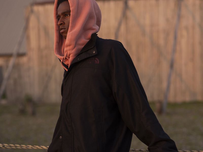 A young adult Black man in a black jacket and pink hoodie poses outdoors, resting his arms on a rope railing against a blurred backdrop.