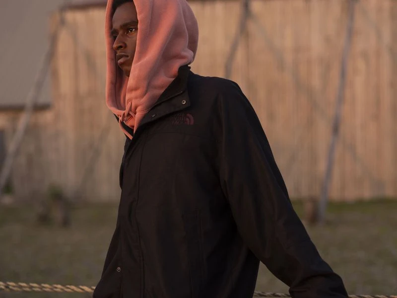 A young adult Black man in a black jacket and pink hoodie poses outdoors, resting his arms on a rope railing against a blurred backdrop.